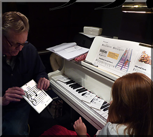 Photo of Jerry Michelsen showing a five year old girl how to read music notes on a staff. They're both seated at the piano.